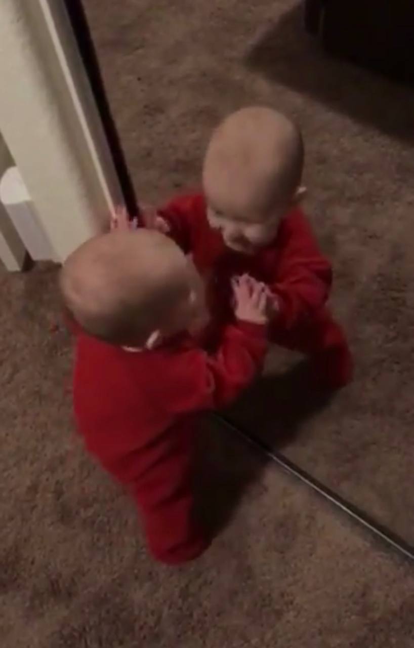 A baby in a red onesie sitting on the floor, both hands pressed against a mirror, face-to-face with his own reflection